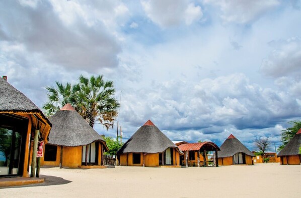 A row of thatched-roof huts with a sandy ground and palm trees in the background. - Ongula Village Homestead Lodge (Ondangwa)