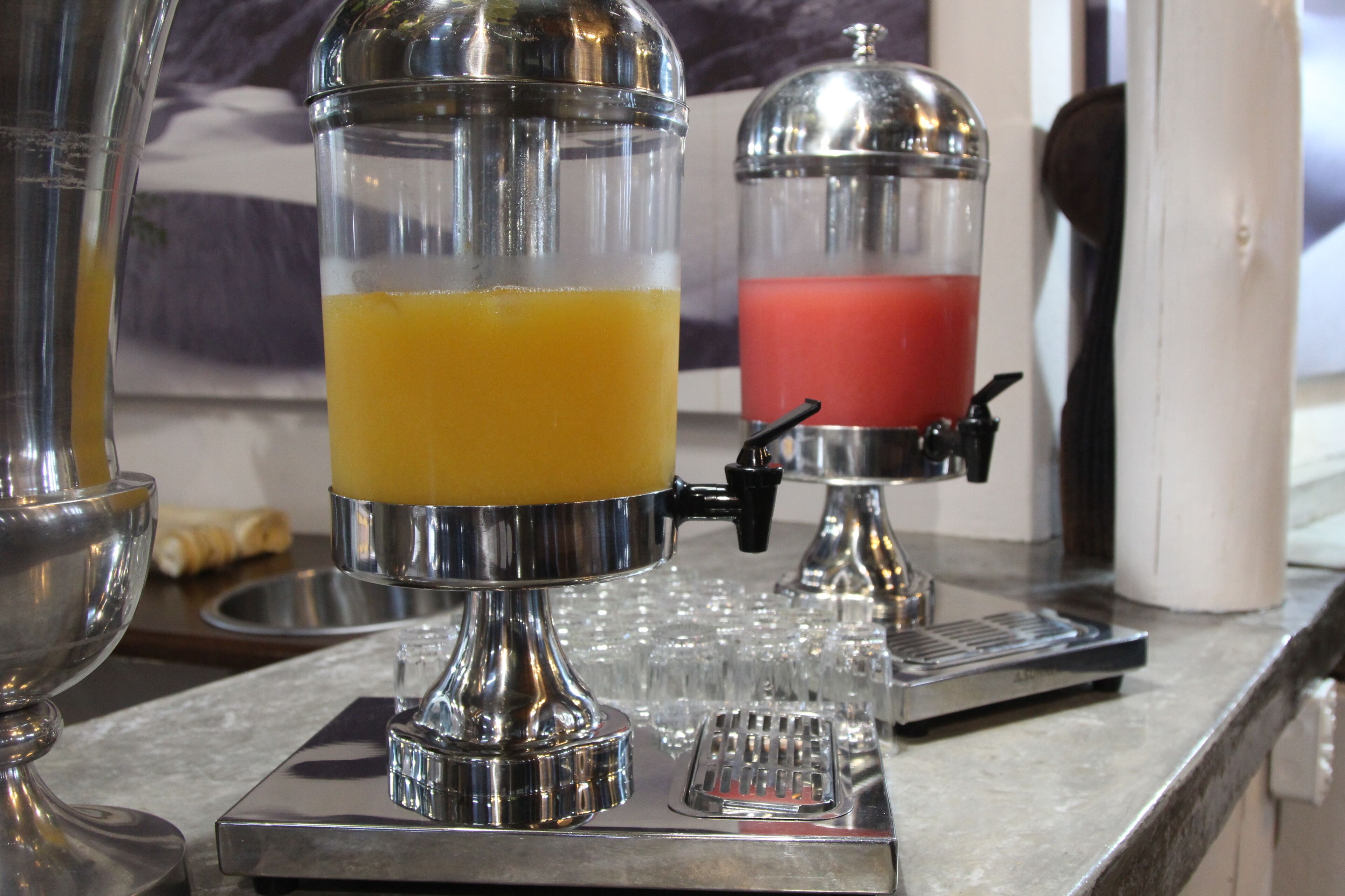 A beverage dispenser with a clear container and a red liquid, a metal bowl, and a tray with glasses.