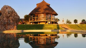 A thatched-roof building with a wooden deck, reflecting in a calm water body. - Lapa Lange Game Lodge (Mariental)