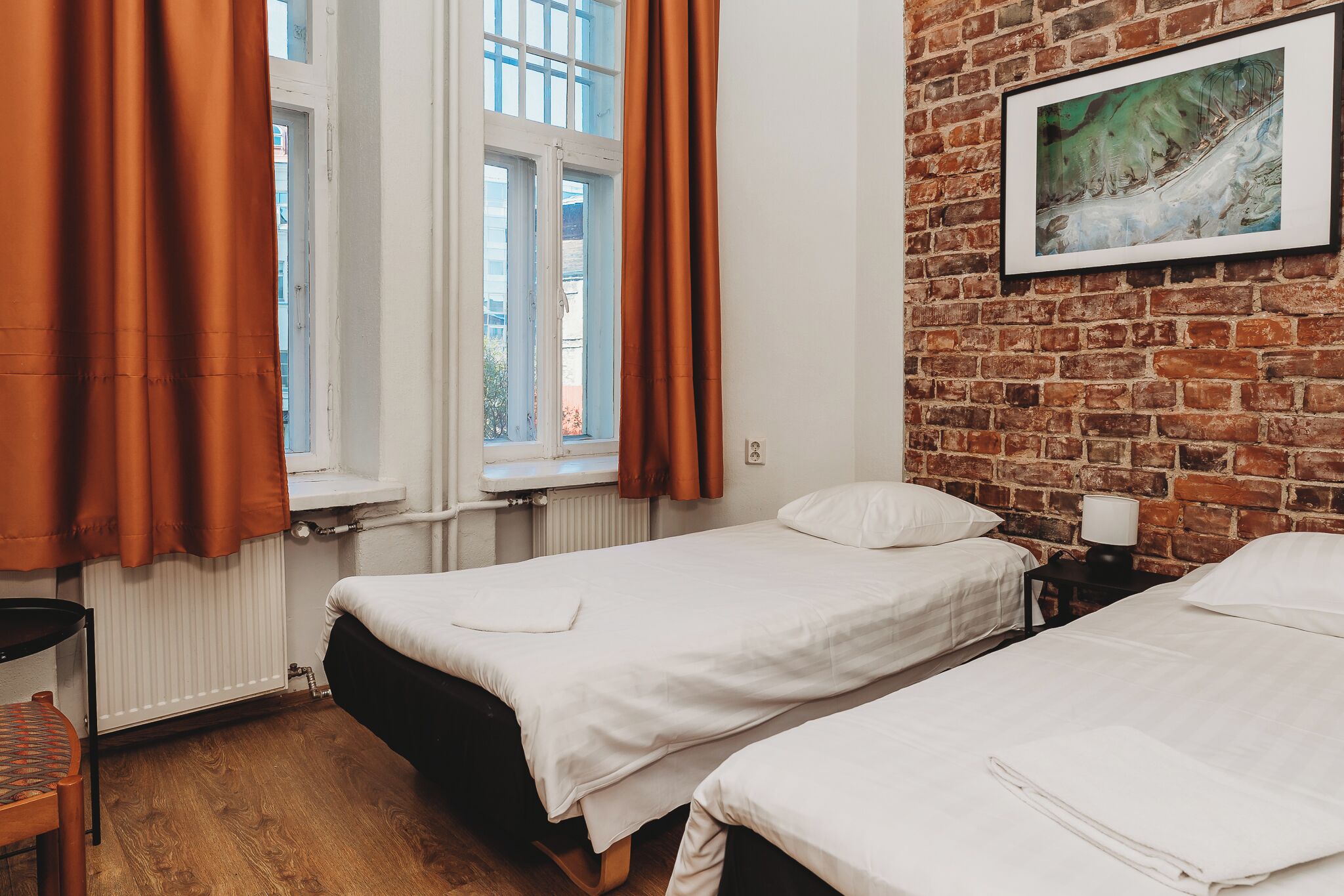 A room with two beds, a brick wall, a window with curtains, a radiator, and a framed picture on the wall.