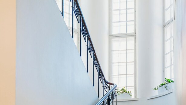 A grand staircase with ornate black railings and marble steps, leading to a landing with large windows and potted plants. - Fat Margaret's Hostel (Tallinn)