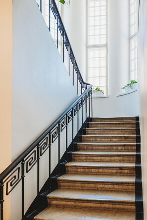 A grand staircase with ornate black railings and marble steps, leading to a landing with large windows and potted plants. - Fat Margaret's Hostel (Tallinn)