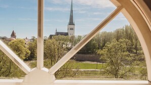 A view through a window with a white frame, featuring a church with a tall spire and a clock tower, surrounded by trees and a grassy area. - Fat Margaret's Hostel (Tallinn)