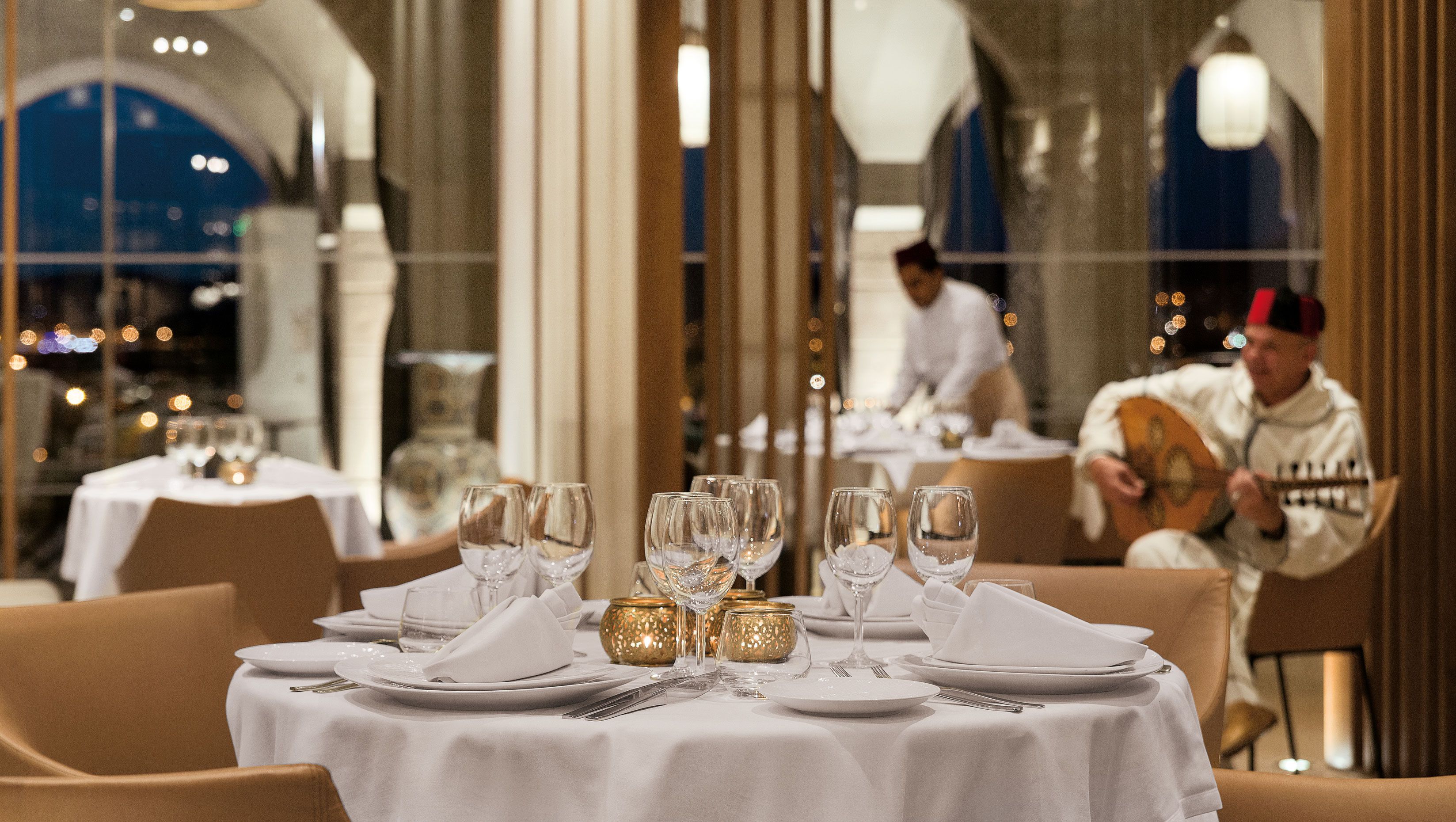 Un ambiente de restaurante con mesas dispuestas para comer, que incluyen manteles blancos, cristalería y candelabros con detalles dorados.