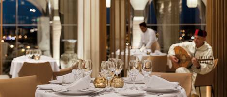 A restaurant setting with tables set for dining, featuring white tablecloths, glassware, and gold-rimmed candle holders.
