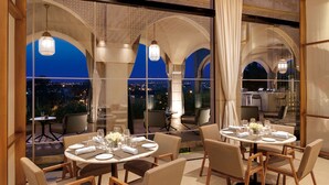 A dining area with round tables set for two, featuring white tablecloths, wine glasses, and cutlery. The space is adorned with hanging light fixtures and has a view of a cityscape at dusk.
