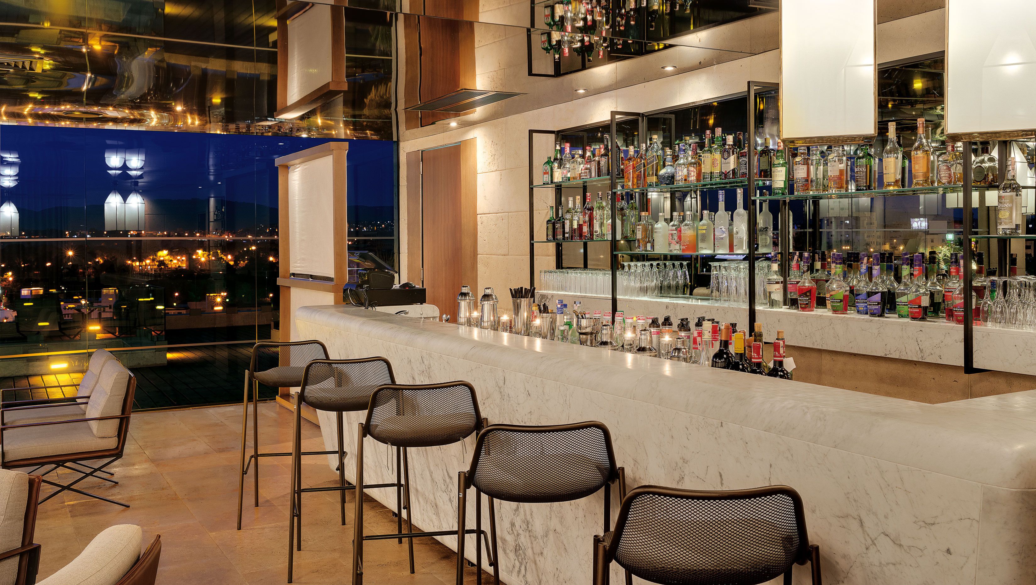Uma área de bar com vista para a cidade, banquetas e uma variedade de garrafas de bebidas.