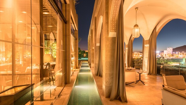 A corridor with a reflective floor, glass walls, and a view of a cityscape at night.