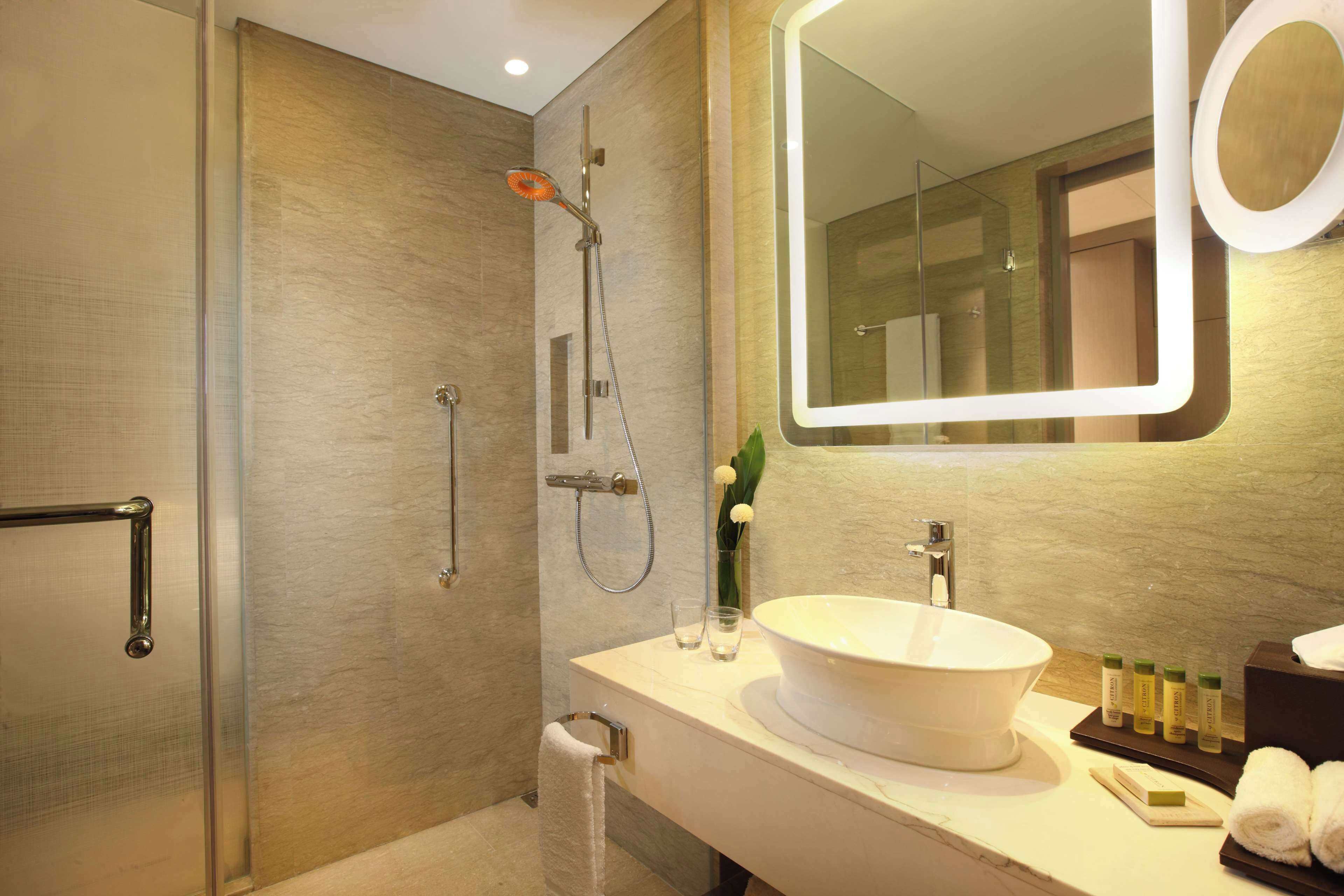 A modern bathroom with a glass shower enclosure, a white sink with a large basin, and a mirror with a circular frame.
