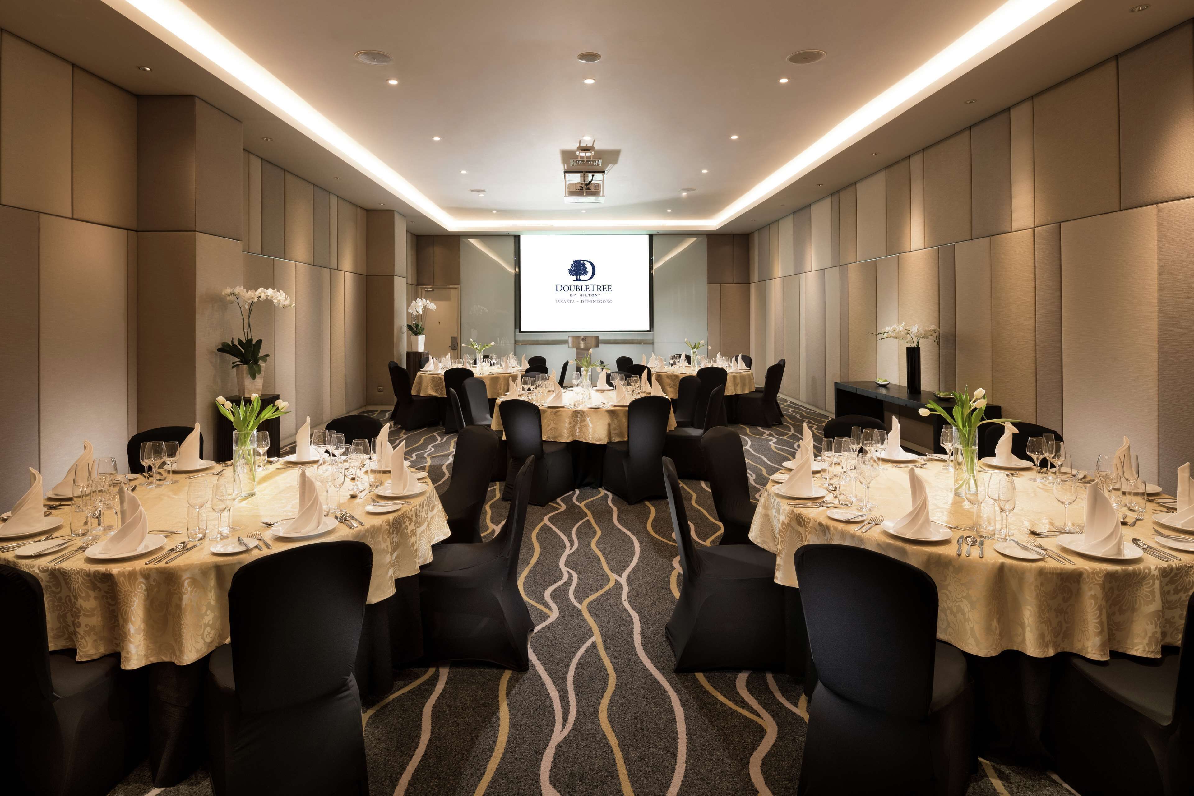 Un salón de conferencias con varias mesas dispuestas para un evento formal, con una pantalla de proyector mostrando el logotipo de DoubleTree.