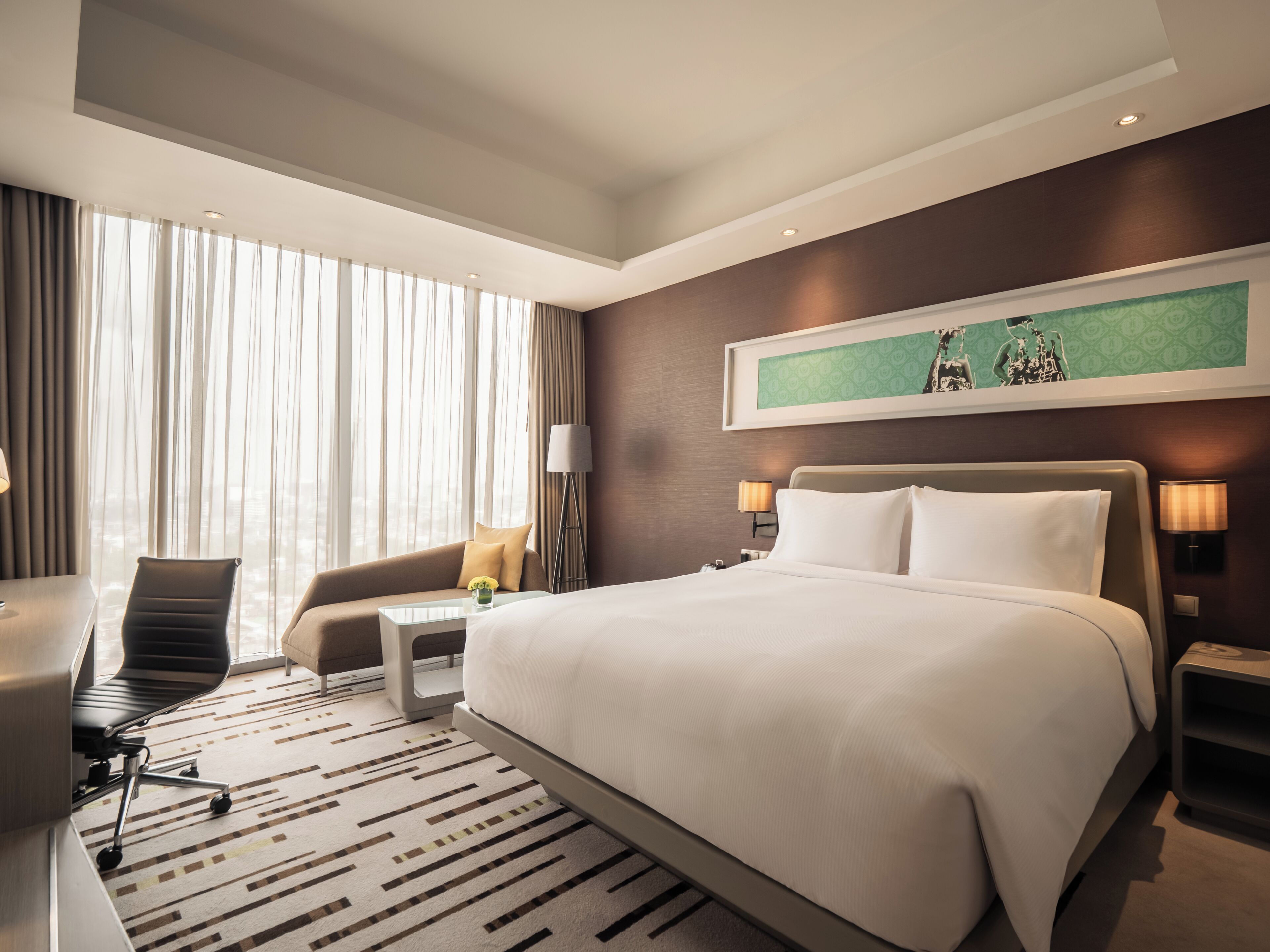 Una habitación de hotel moderna con una cama grande, un escritorio, una silla y un sofá. Hay dos lámparas de noche y un ventanal con cortinas.
