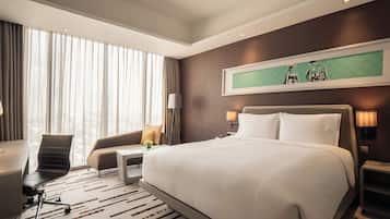 Una habitación de hotel moderna con una cama grande, un escritorio, una silla y un sofá. Hay dos lámparas de noche y un ventanal con cortinas.