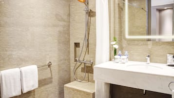 A bathroom with a marble wall, a shower with a handheld showerhead, a white sink with a mirror, and a towel rack with two towels.