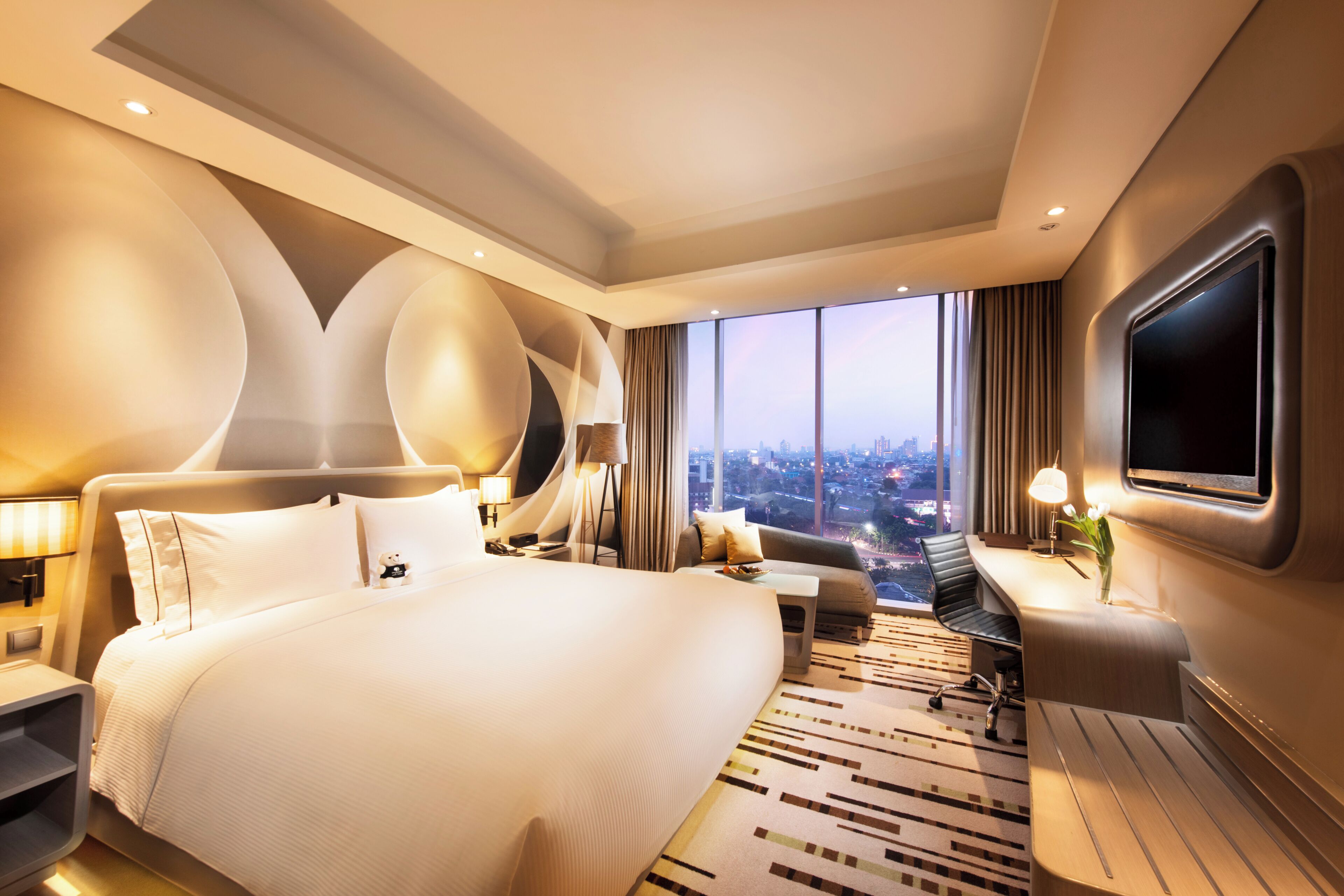 Una habitación de hotel moderna con una cama grande, un escritorio, una silla, un televisor y vistas a la ciudad.
