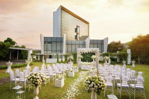 An outdoor wedding venue with white chairs arranged in rows, a floral arch, and a modern building in the background. - DoubleTree by Hilton Jakarta - Diponegoro (Jakarta)
