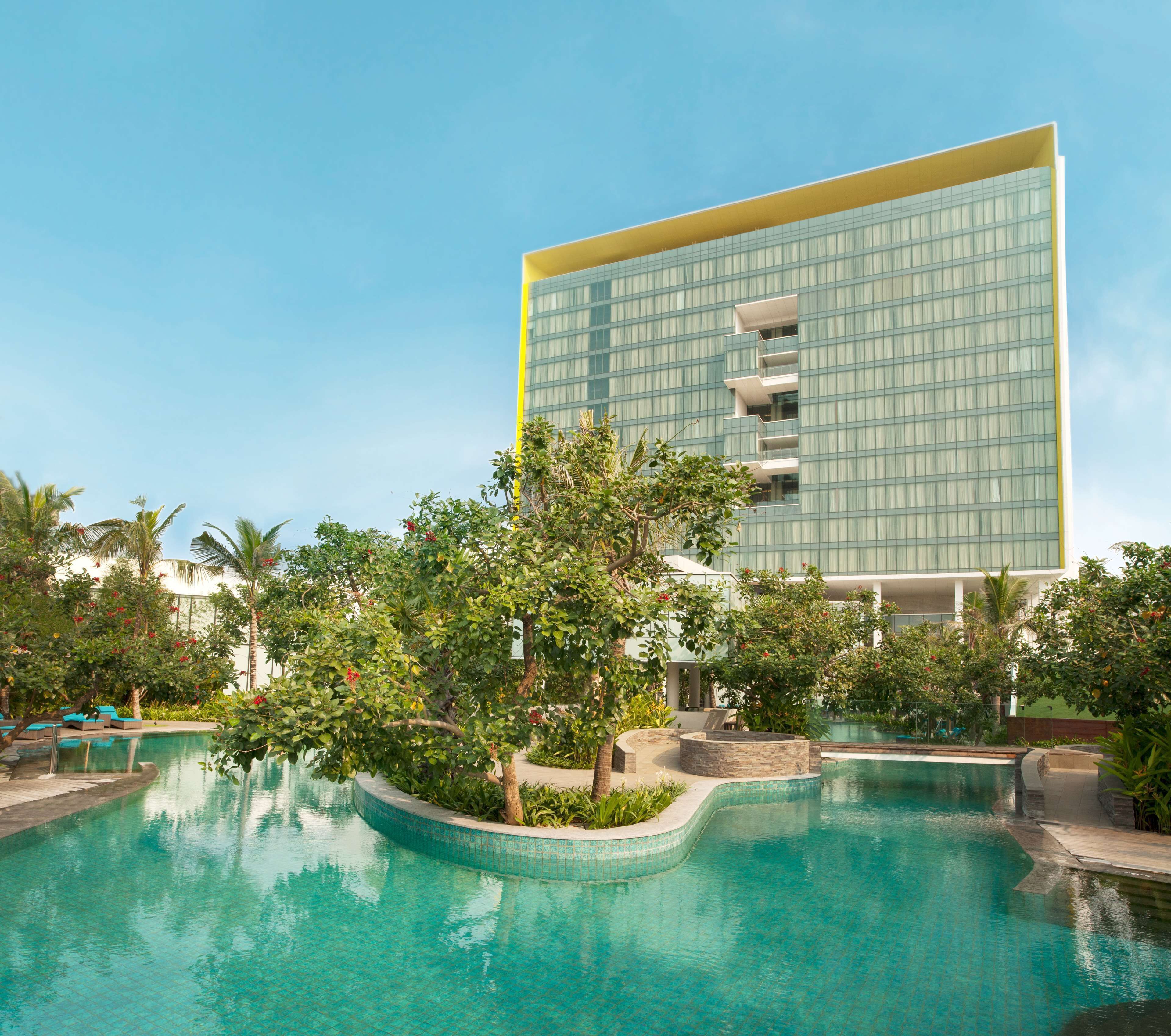 Un hotel moderno con un amplio frente de vidrio, piscina y abundante vegetación.