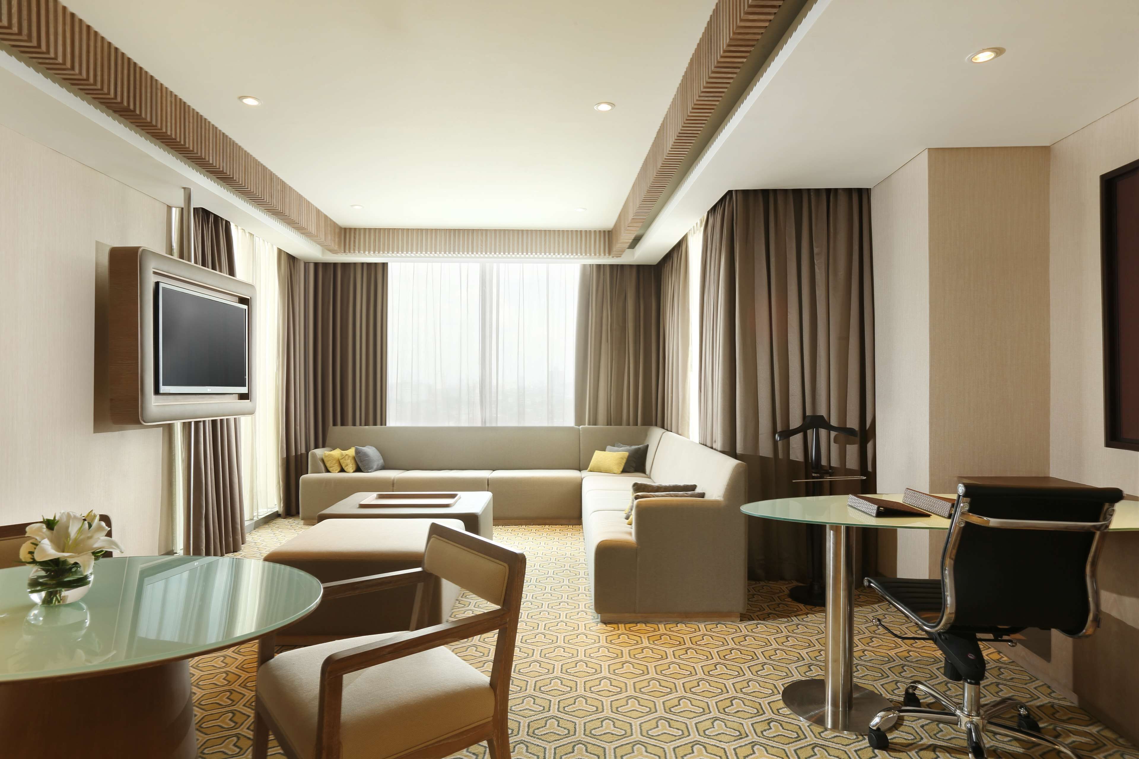A hotel room with a sofa, a glass dining table, a TV mounted on the wall, and a desk with a chair.
