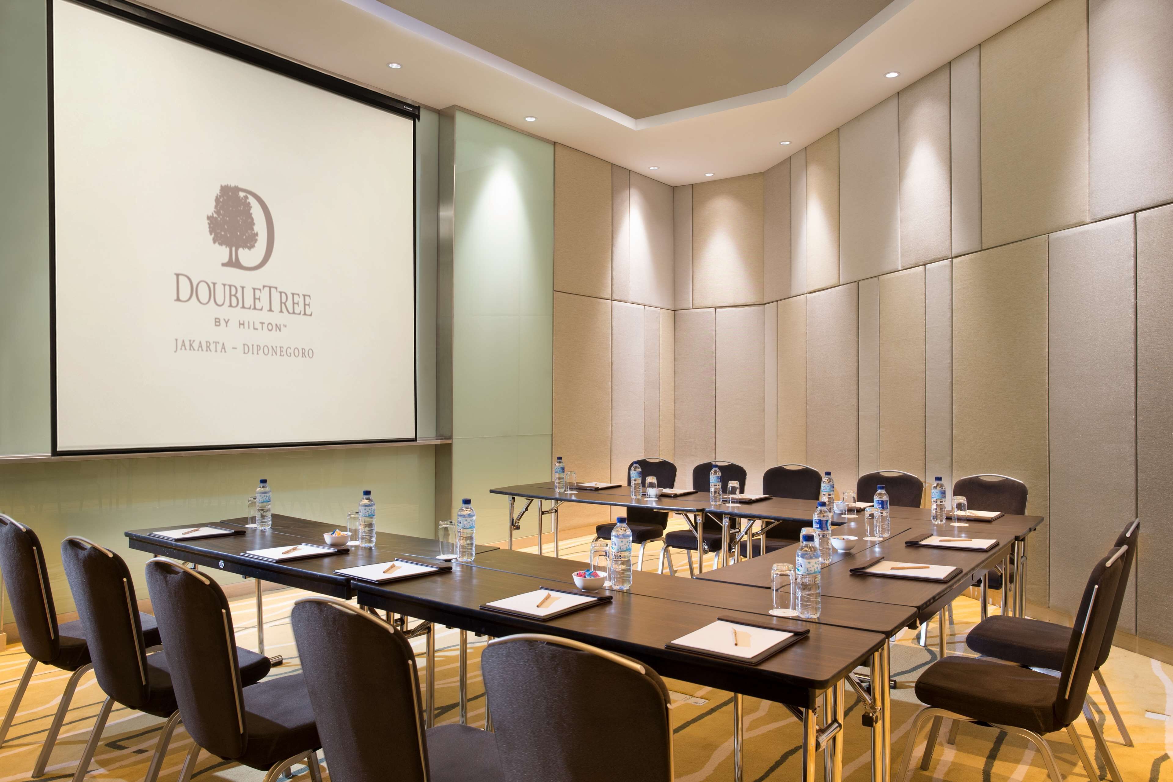Un salón de conferencias con una larga mesa, sillas y una pantalla proyectora mostrando el logotipo de DoubleTree Jakarta.