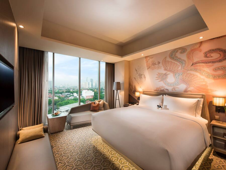 A modern hotel room with a large bed, a sofa, a TV, and a city view.