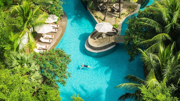 Vista aérea de un complejo turístico con una piscina central, rodeado de palmeras y zonas de descanso.