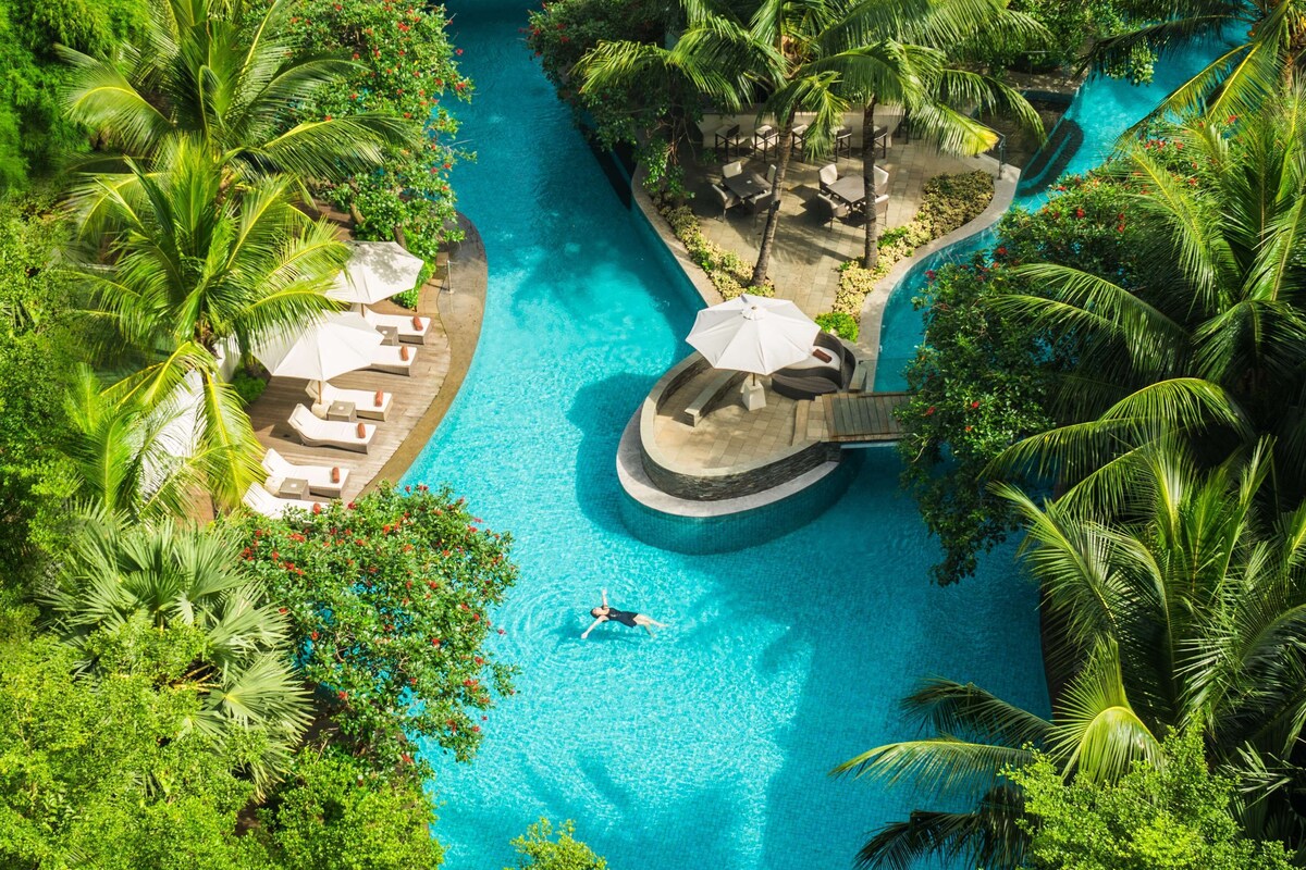 An aerial view of a resort with a central swimming pool, surrounded by palm trees and lounge areas.