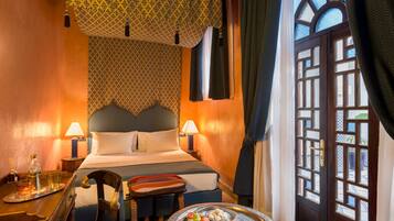 A hotel room with a bed, bedside lamps, a tray with fruits, and a view through a window with patterned curtains.