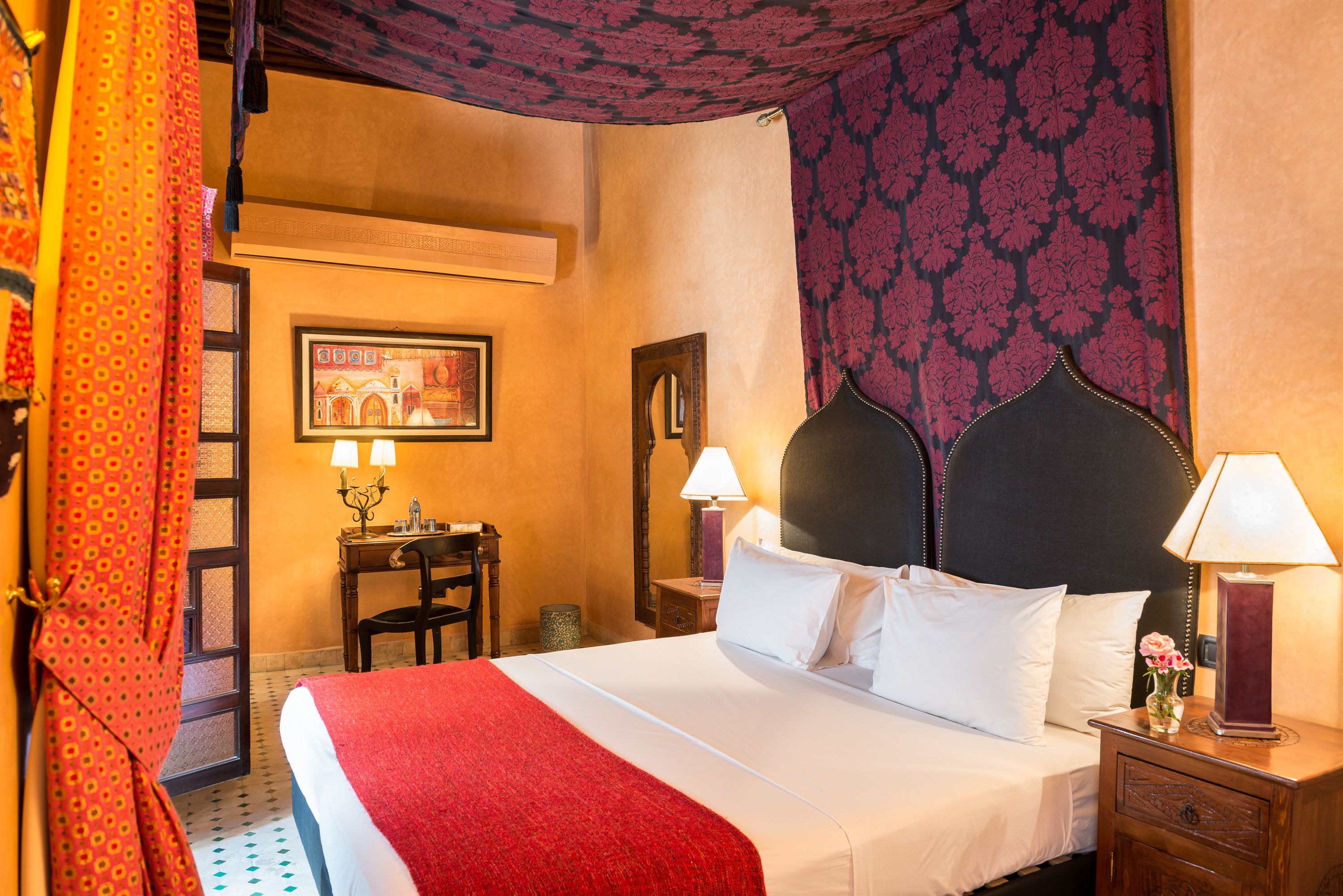 Quarto de hotel com cama, escrivaninha e cadeira. Há uma janela com cortinas, uma luminária na escrivaninha e outra na mesa de cabeceira.