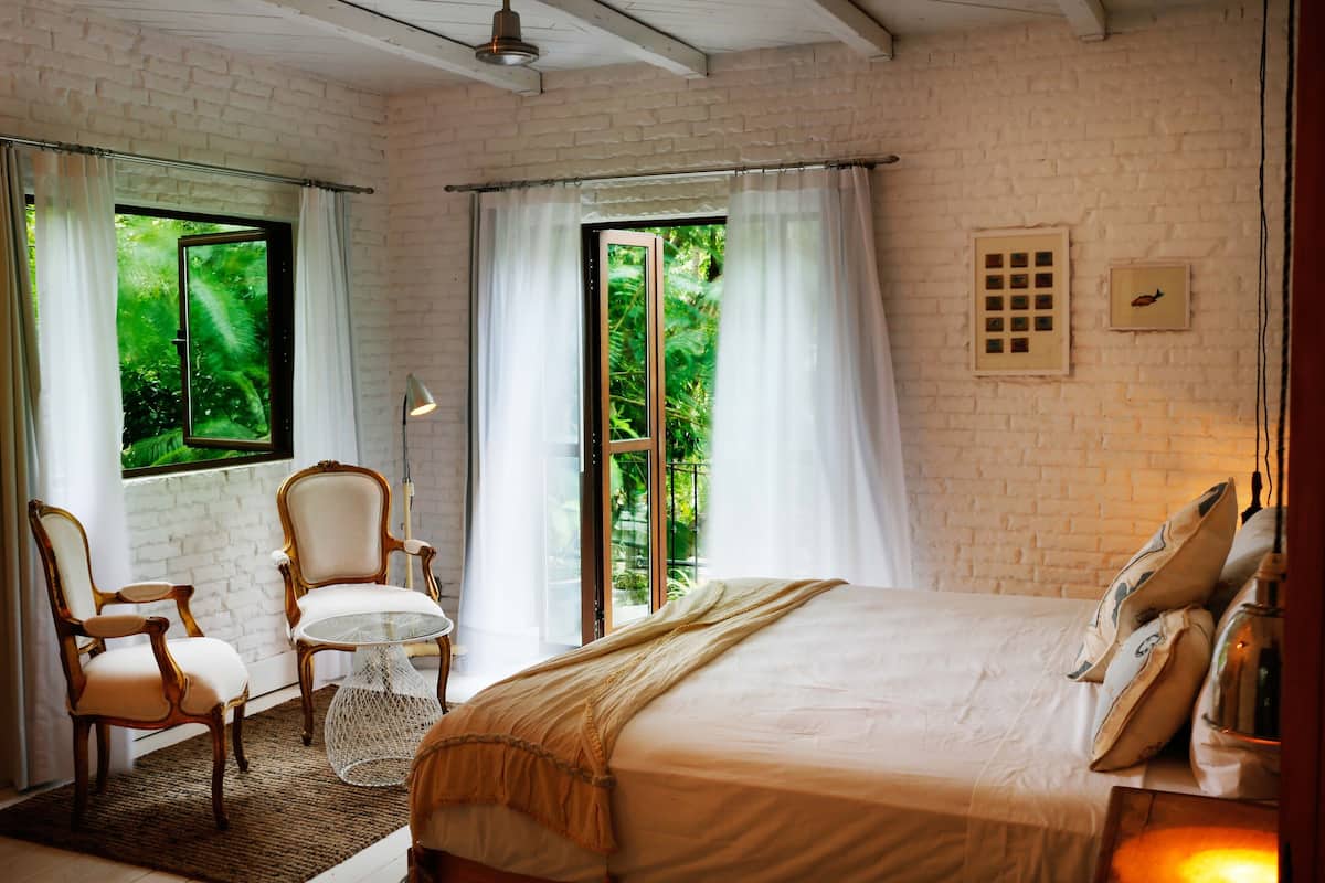 A bedroom with a large bed, two chairs, a small table, and a window with curtains.
