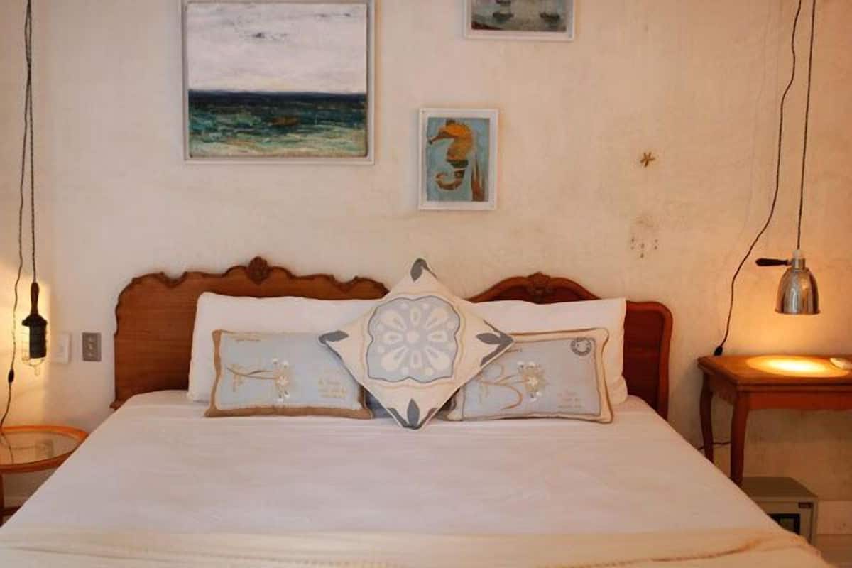 A bedroom with a wooden headboard, a bed with white bedding, and two pillows with decorative patterns. There are two framed artworks on the wall and a small table with a lamp beside the bed.