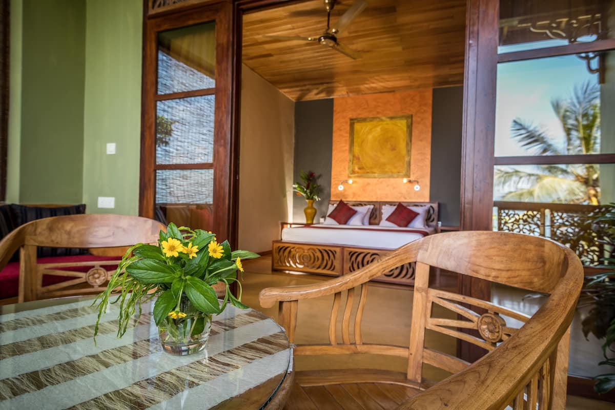 A wooden-framed room with a bed, a table with a vase of flowers, and a view of palm trees through large windows.