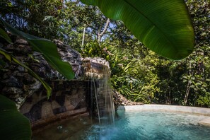 Uma cascata desemboca em uma piscina rodeada por vegetação exuberante e um muro de pedra.