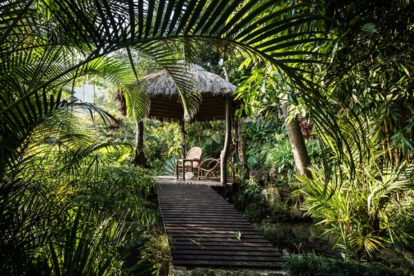 A wooden pathway leading to a thatched-roof structure surrounded by lush greenery.