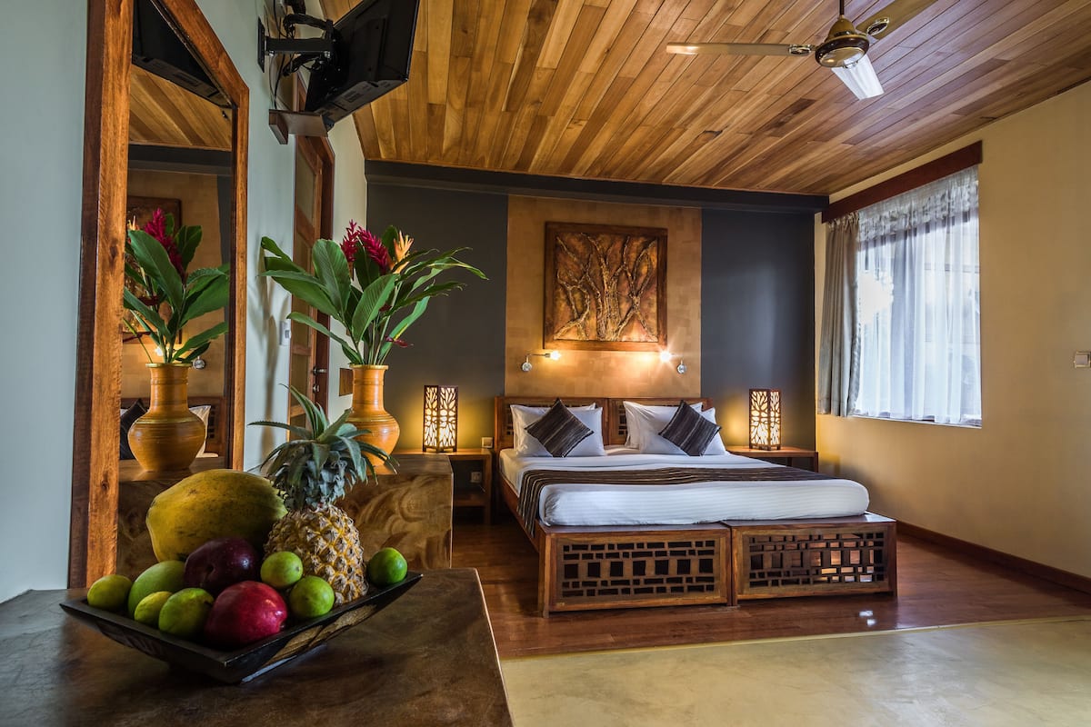 A bedroom with a wooden ceiling, a bed with white bedding, a fruit bowl on a table, and a window with curtains.