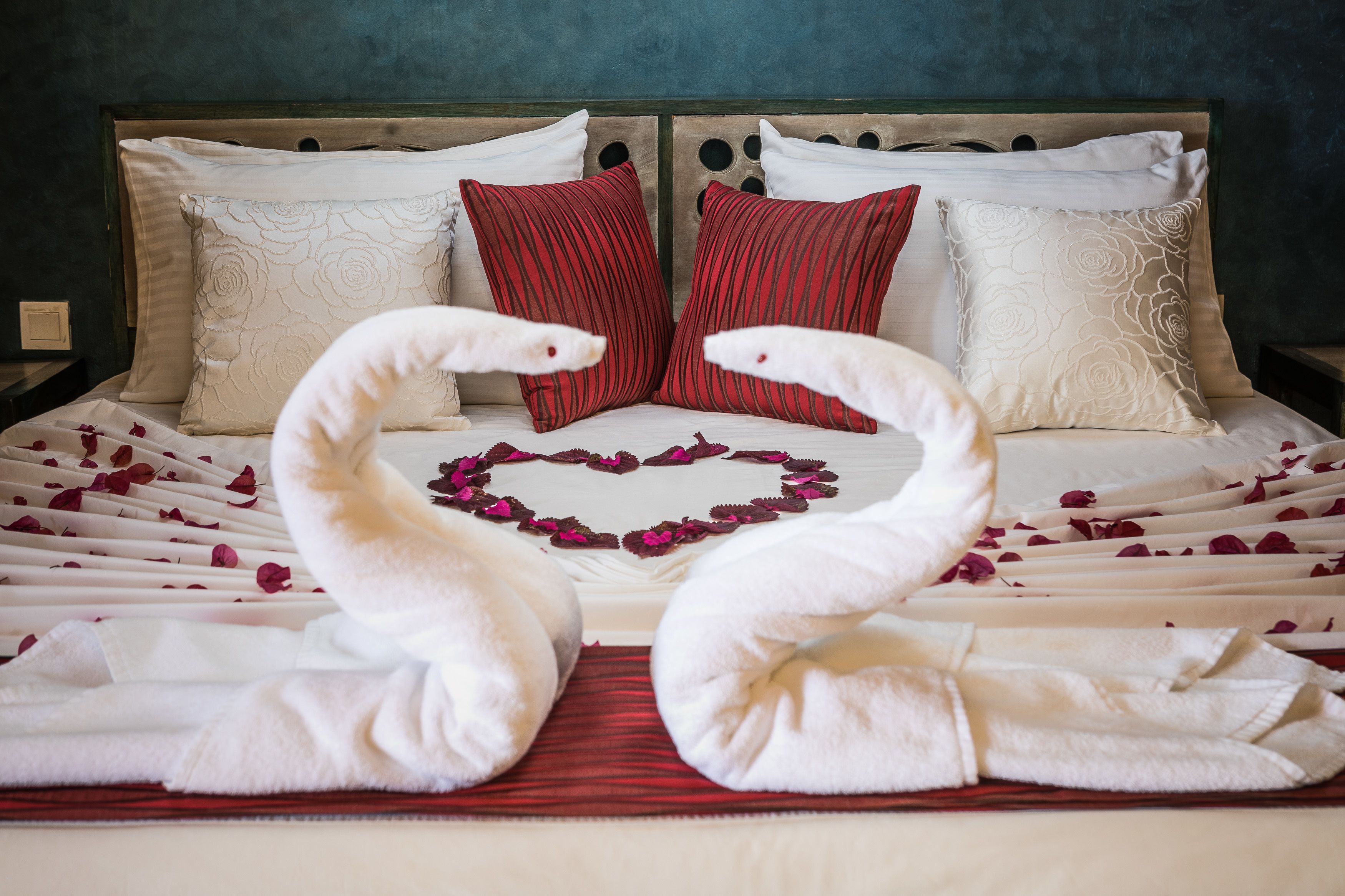 A bed with white linens, red and white pillows, and a heart-shaped arrangement of rose petals.