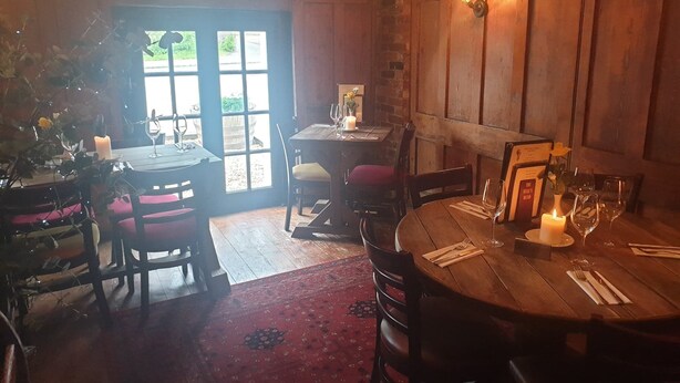 A cozy dining area with wooden furniture and a red carpet. There are two tables, one round and one rectangular, both set with wine glasses and candles. - The Bulls Head (Chichester)
