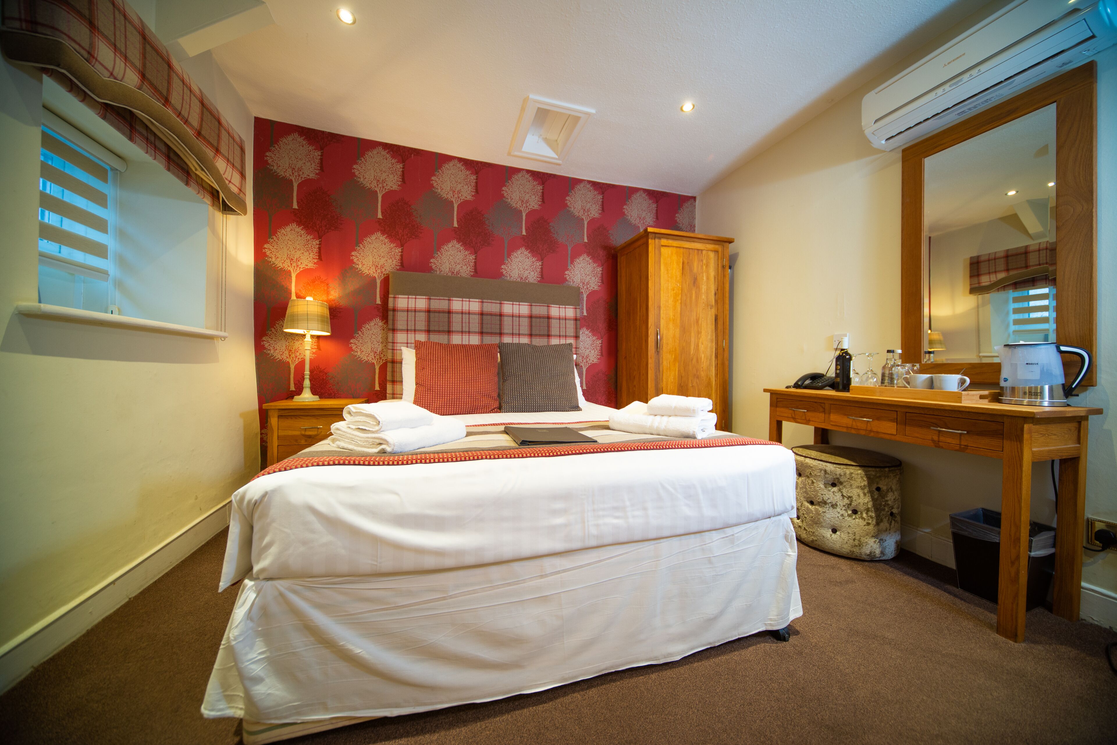A neatly made bed with white linens, a wooden wardrobe, a desk with a mirror, and a red patterned wall.
