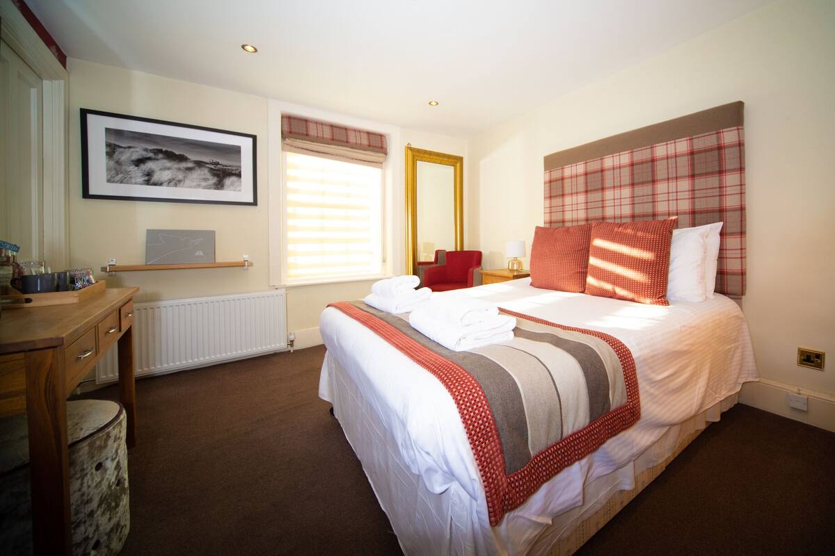 Een hotelkamer met een bed, een bureau, een stoel, een raam met jaloezieën en een ingelijst beeld aan de muur.