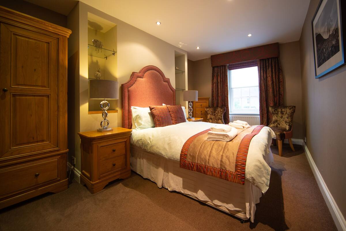 Een goed verlichte slaapkamer met een groot bed, een nachtkastje, een dressoir en een raam met gordijnen.
