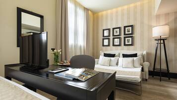A hotel room with a desk, a bed, a chair, a TV, a lamp, and framed pictures on the wall.