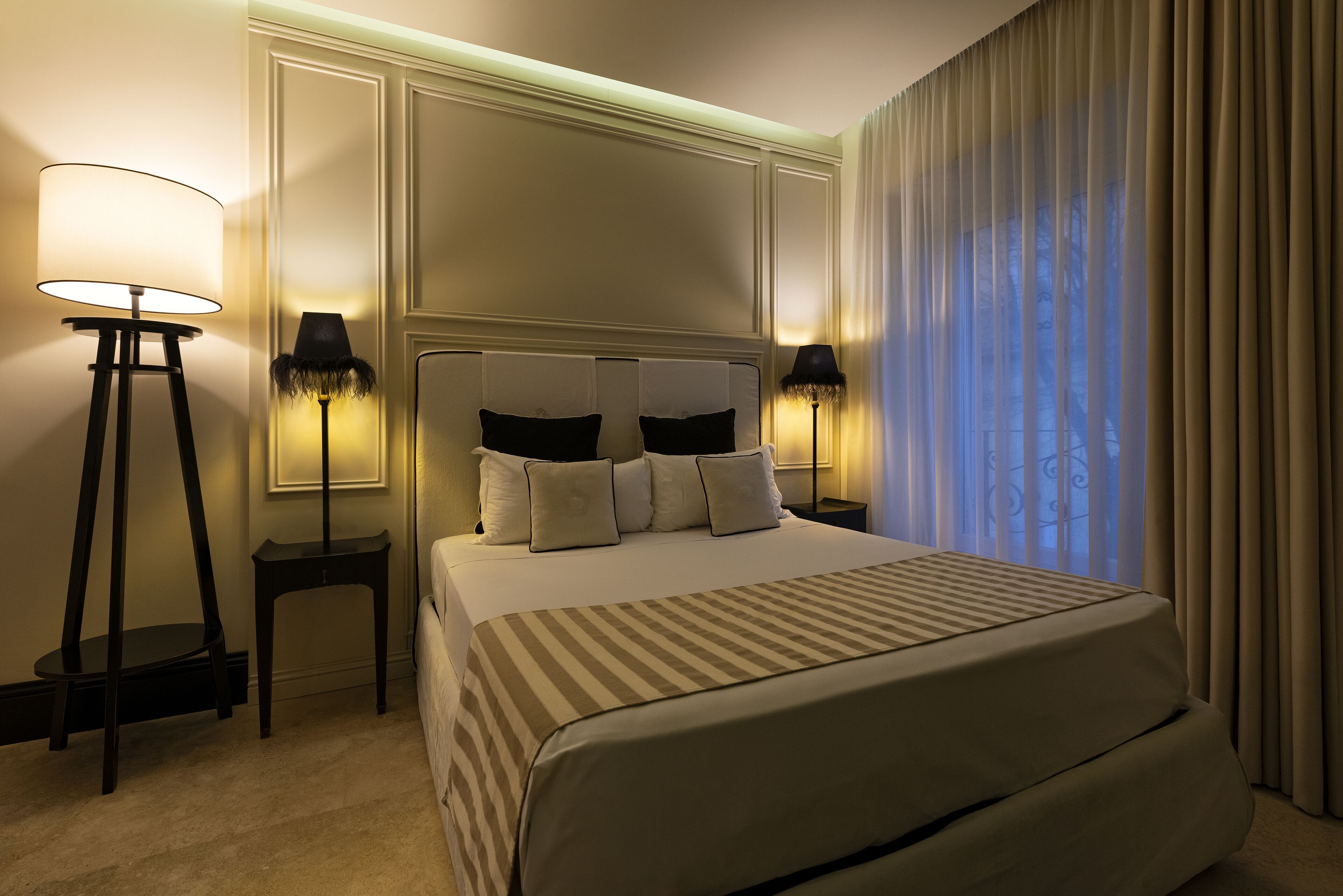A hotel room with a large bed, two bedside lamps, a headboard with a mirror, and a view through sheer curtains.