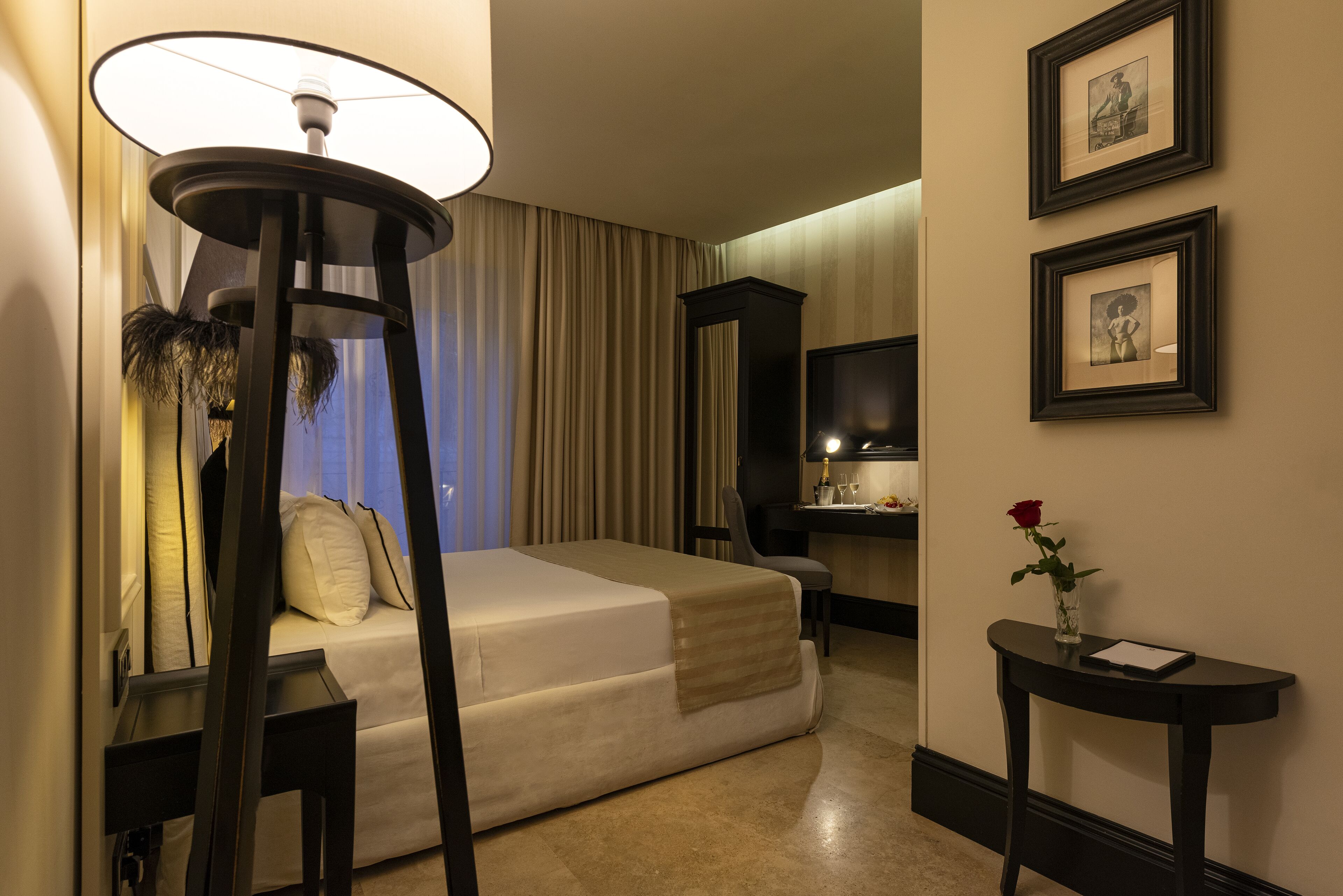 A hotel room with a bed, a desk, a chair, a lamp, a vase with a rose, and framed pictures on the wall.