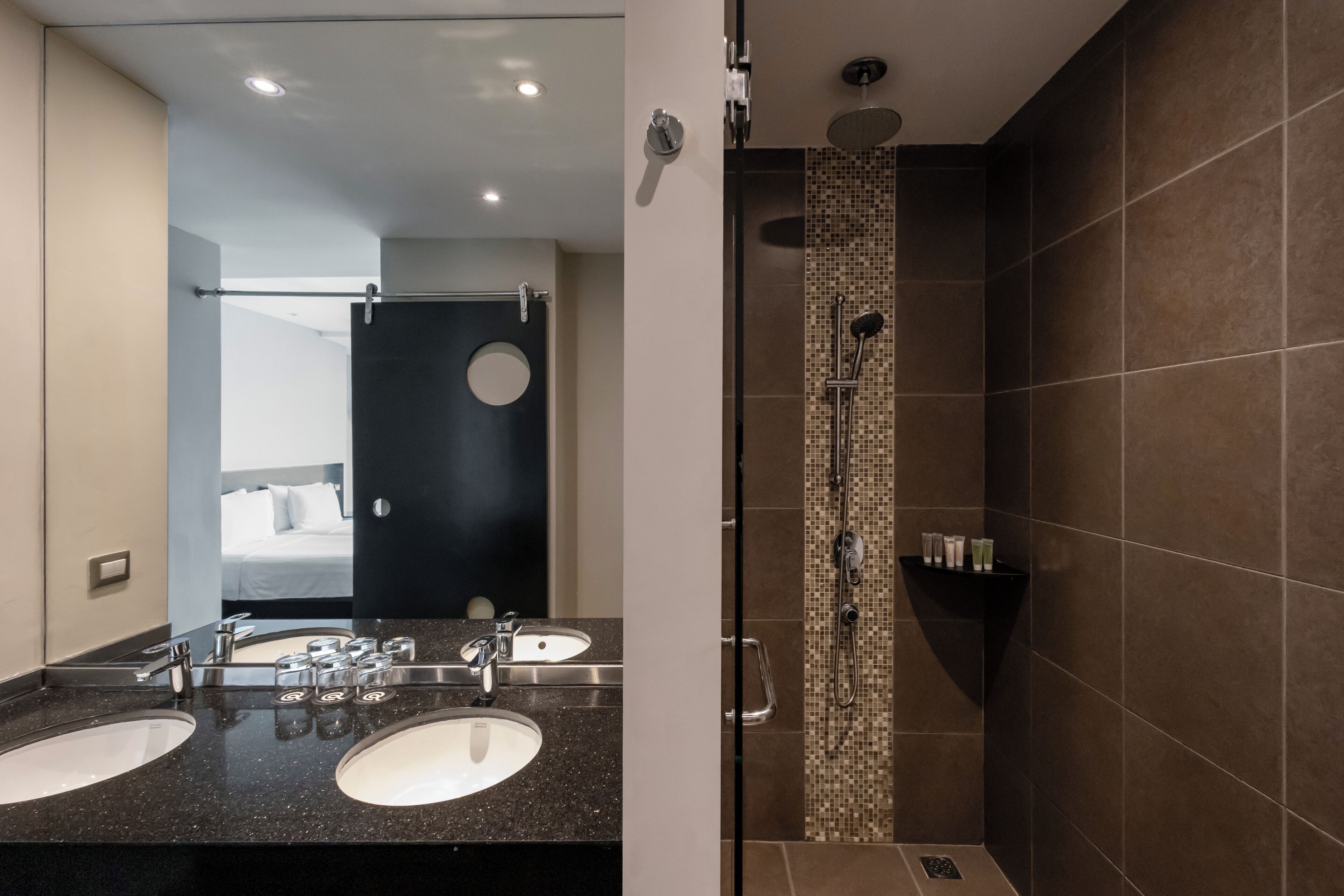 Une salle de bain moderne avec deux lavabos, une douche et un miroir qui reflète une chambre à coucher.