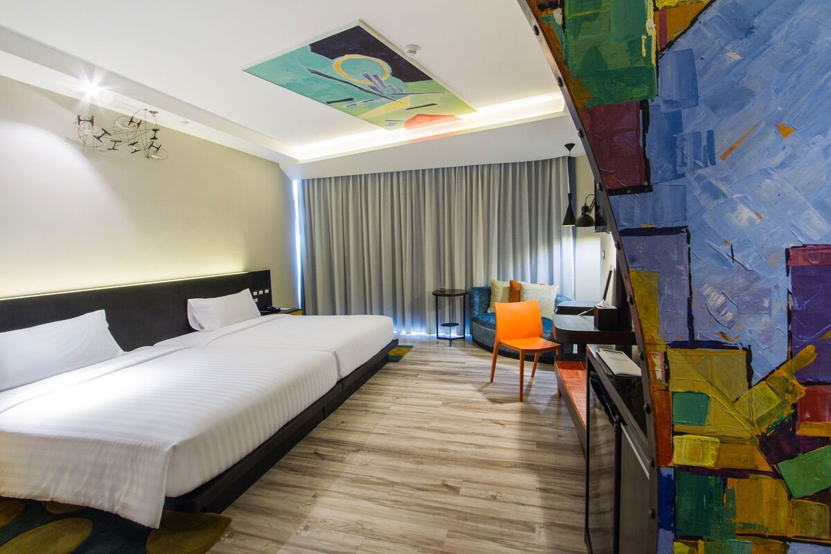 A hotel room with a bed, a desk, a chair, a desk lamp, and a colorful abstract painting on the wall.