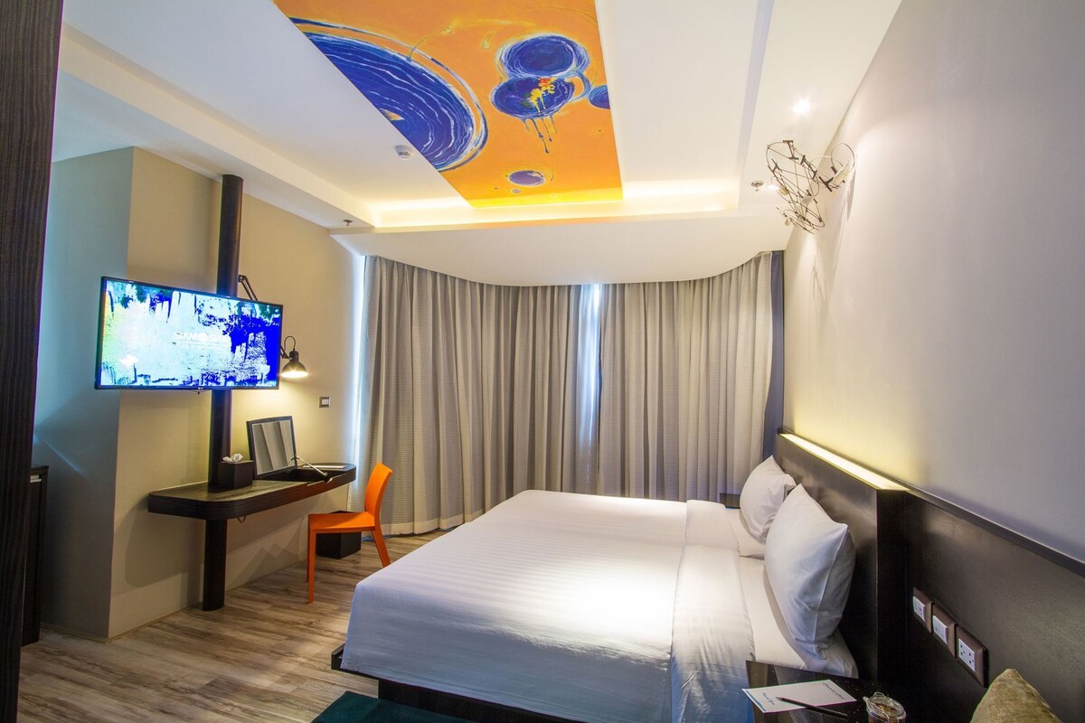 A hotel room with a bed, a desk with a TV, a chair, and a ceiling with a unique design.