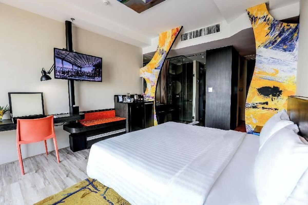 A modern hotel room with a large bed, a TV mounted on the wall, a desk with a chair, and a wardrobe.