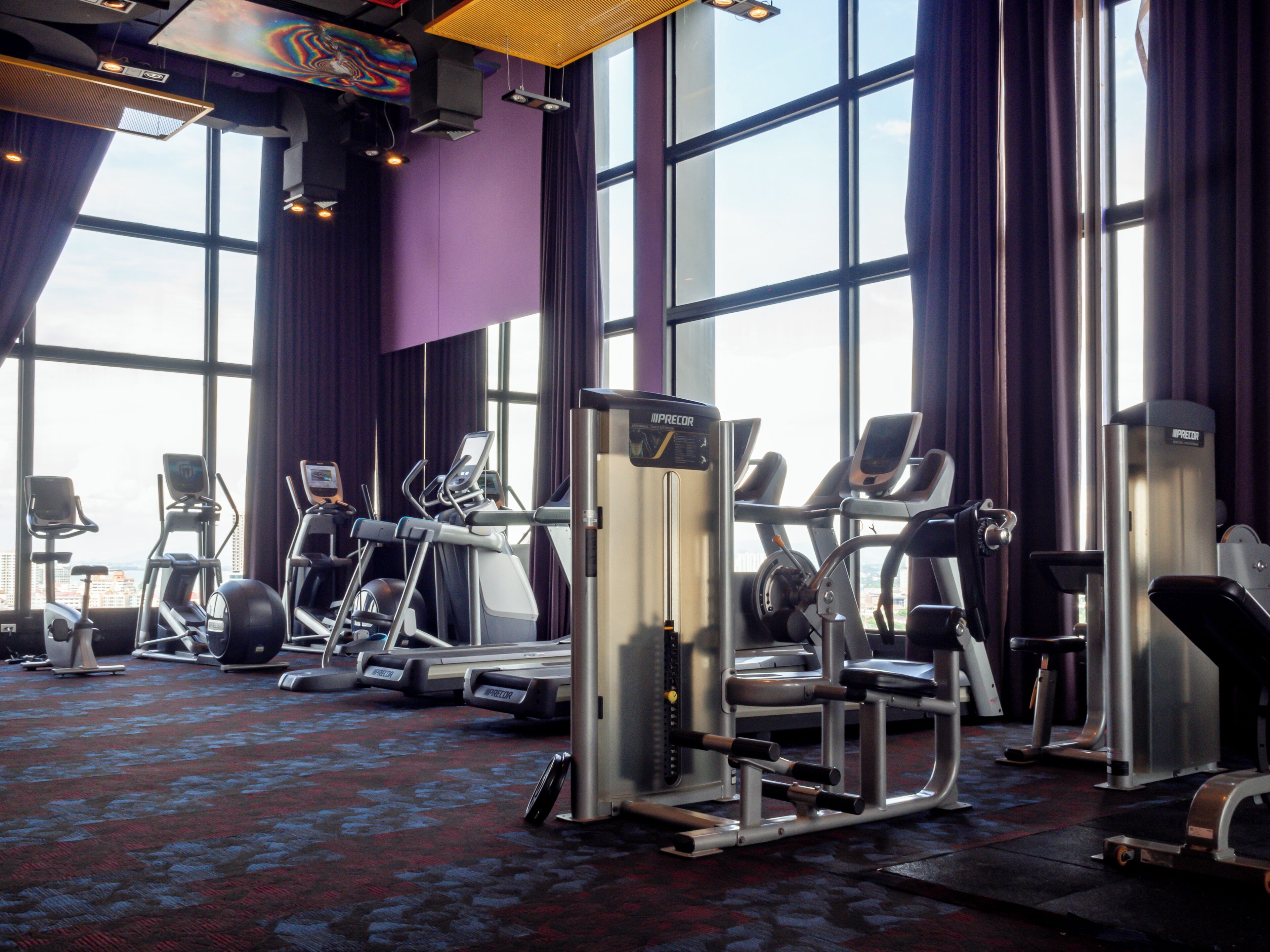 A gym with various exercise machines, large windows, and purple walls.