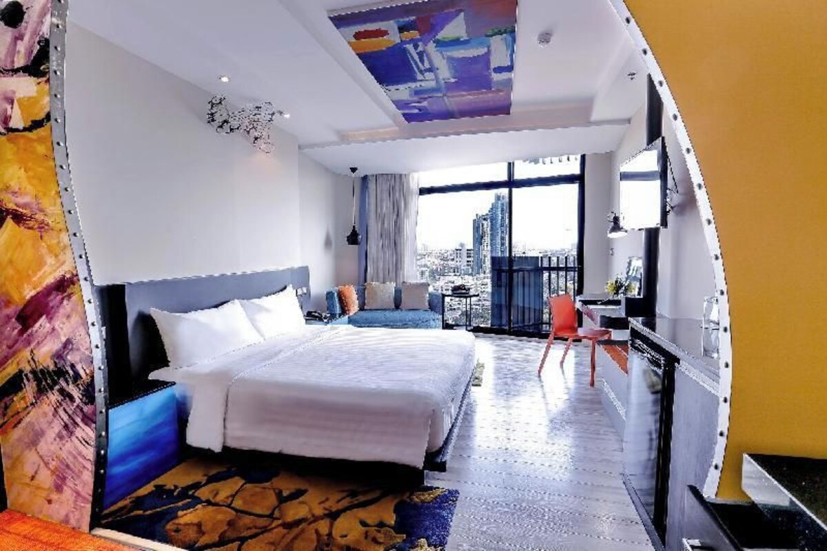 A modern hotel room with a large bed, a seating area, a TV, and a balcony with a city view.