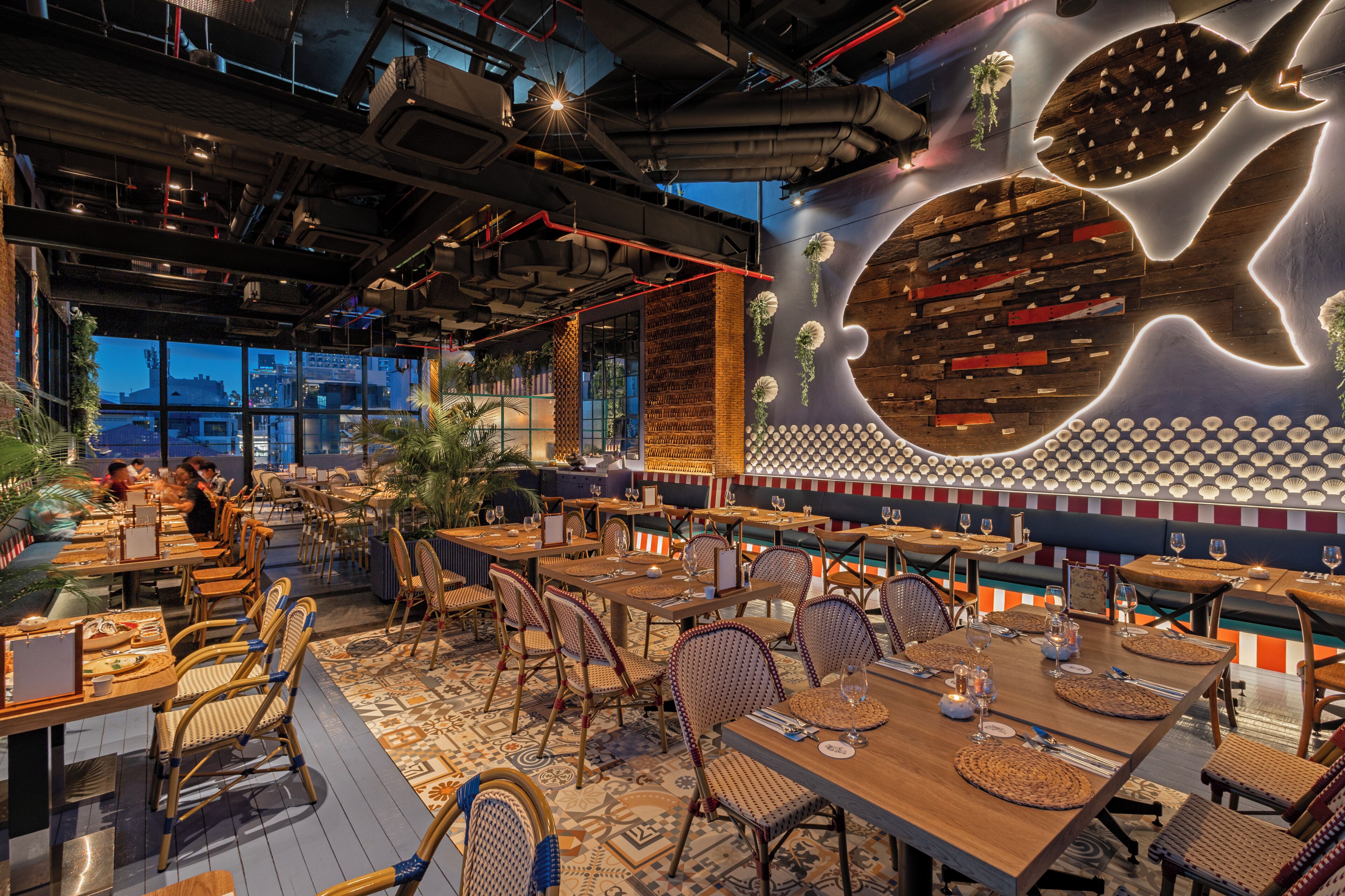 A restaurant with multiple dining tables and chairs, a large illuminated wall art piece, and a view of the cityscape through large windows.