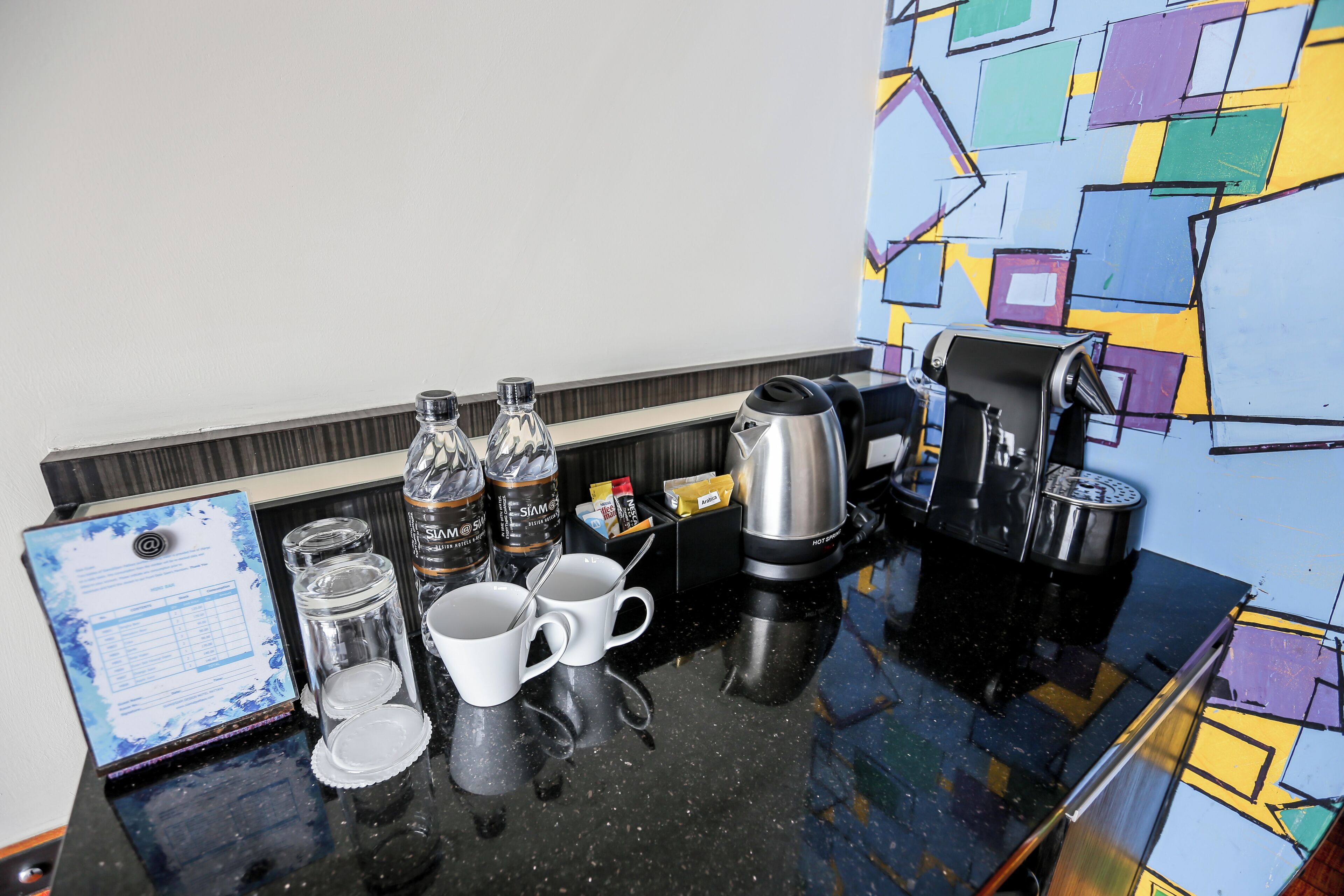 A hotel room's breakfast area with a coffee maker, two cups, a water bottle, and a menu on the counter.
