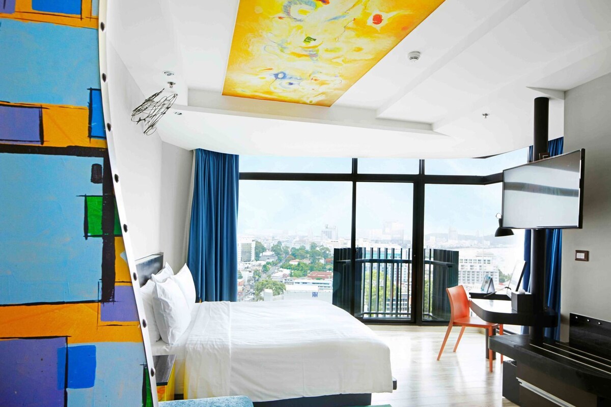 A hotel room with a bed, a TV, a desk, and a large window with a city view.